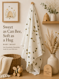 A soft white baby wrap blanket with a scattered yellow and black bumblebee print hangs from a wooden peg on a beige wall. The scene is styled as a cozy nursery with a framed beehive illustration, a wooden toy bee, a woven basket with a cream knit throw, a wooden honeycomb shelf, and a ribbed cream vase with dried grasses.