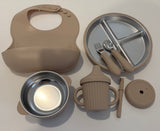 A baby feeding set in oatmeal and stainless steel, including a silicone bib, divided plate with utensils, a bowl, a handled cup, a lid, and a small brush, arranged on a light gray background.