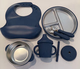 A baby feeding set arranged on a light gray background, including a navy blue silicone bib, a divided stainless steel plate with matching utensils, a small stainless steel bowl, a navy blue handled cup, a lid, and a small cylindrical accessory.