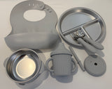 A baby feeding set in matte gray and metallic silver, including a silicone bib, divided plate, bowl, sippy cup, spoon, brush, and lid, arranged on a white background.
