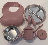 A baby feeding set in pink and stainless steel, including a silicone bib, divided plate with utensils, a bowl, a handled sippy cup, a straw, and a lid, arranged on a white background.