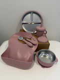 A pink baby feeding set arranged on a white surface against a light gray wall, including a silicone bib, spoon and fork, a sippy cup with straw, a divided plate, and a bowl with lid.