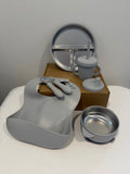 Gray silicone baby feeding set including a bib, spoon, fork, sippy cup with straw, bowl, and a three-compartment plate, arranged on a white surface with a brown box in the background.