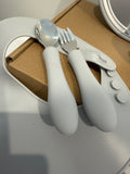 A baby utensil set with white silicone handles and silver metal heads, including a spoon and a fork, resting on a white silicone holder over a brown cardboard box.