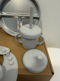 A light gray silicone baby tummy cup with a lid, straw, and side handles, shown alongside its matching lid and a stainless steel divided plate on a cardboard box.
