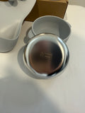 A stainless steel round bowl or plate shown open with its gray lid resting beside it on a white surface, with a brown box partially visible in the background.