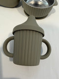 A ribbed silicone baby cup with dual side handles and a spout, shown in a muted taupe-gray color on a white surface. Another matching cup and a stainless steel bowl are partially visible in the background.