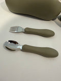 A child's silicone spork and spoon set with olive green handles and silver stainless steel bowls, displayed on a white surface with a matching case in the background.