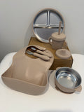 A baby feeding set in a warm beige/tan color, including a silicone bib with integrated utensils, a stainless steel divided plate, a sippy cup with straw, a small bowl, and a lid, arranged on a white surface with its cardboard box in the background.