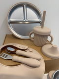 A coordinated baby feeding set arranged on a cardboard box against a plain wall. The set includes a round divided metal plate, a ribbed beige sippy cup with a straw and matching lid, a beige silicone bib, a silicone spoon cover, and utensils with beige handles and silver metal heads.