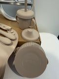 A beige silicone baby feeding set including a plate, spoon, cup with straw, lid, and bowl, shown on a white surface with a cardboard box in the background.