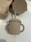 A beige silicone baby sippy cup with dual handles and a straw, shown alongside matching silicone bowls and a plate on a white surface.