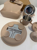 A multi-piece baby feeding set in a warm beige silicone and stainless steel color combination, including a silicone bib, plate, cup, and accessories, arranged on a white surface.