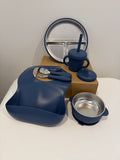 A baby feeding set in navy blue and stainless steel, including a silicone bib, sippy cup with straw, bowl, spoon, and a plate, arranged on a white surface with a brown box in the background.