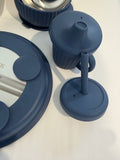 A set of matte navy blue silicone baby feeding accessories, including a cup with a spout and handle, a round base, and a lid, arranged on a white surface.