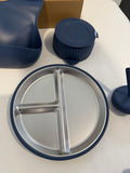 A stainless steel divided baby plate with a navy blue rim, shown alongside matching navy blue accessories including a silicone cup, a small container, and a stand on a white surface.