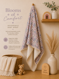 A reversible baby wrap blanket with a pastel floral print on one side and a cream muslin reverse, hanging on a wooden wall hook in a styled nursery setting. The image includes promotional text reading 'Blooms of Comfort' and 'Reversible Baby Wrap,' along with props like a basket, teddy bear, shelf, dried grass, and a wooden sign.