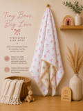A baby wrap blanket with a pink bow and snowflake pattern on a white muslin fabric, hanging on a wooden stand in a styled nursery setting. The image includes promotional text on the left, a basket with a cream knit blanket, a wooden bunny toy, a ceramic vase with dried grass, and a wooden shelf with a small wooden arch and vase.