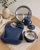 A coordinated children's dinner set arranged on a woven placemat on a light wood table. The set includes a navy blue silicone bib, a matching navy cup with straw, a stainless steel divided plate, a navy bowl, a lid, utensils, and a small vase with greenery in the background.