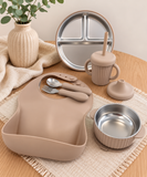 A coordinated baby feeding set arranged on a wooden table, including a beige silicone bib, stainless steel bowl, cup, spoon, lid, and a divided plate, styled with a cream cloth and a vase of greenery.