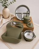 A coordinated baby feeding set in muted olive green and stainless steel, arranged on a neutral fabric surface with a vase and leaves as styling props. The set includes a large silicone bib, a divided plate, a cup with straw, a bowl, utensils, and a small container.