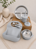 A baby feeding set arranged on a beige fabric surface, including a light gray silicone bib, stainless steel bowl, cup with straw, plate, and utensils, with a brown box in the background and a vase of greenery at the side.