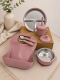 A coordinated baby feeding set in dusty pink and stainless steel, arranged on a beige textile with a box behind it. The set includes a large silicone bib, a divided plate, a sippy cup with straw and lid, a small bowl, and utensils.