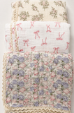 Three folded quilted muslin blankets stacked vertically, each with a different floral and bow pattern. The top blanket has green floral motifs on a cream background, the middle has pink bows on a white background, and the bottom has a pink and lavender floral print with cream lace trim.