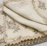 A folded cream-colored cotton muslin quilt with a delicate pink and green floral print and a scalloped lace border, shown on a white background.