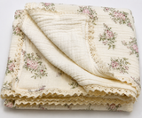 A folded cream-colored cotton muslin blanket with a pink and green floral print and lace trim, shown on a clean white background.
