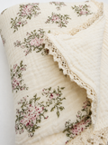 A close-up of a cream-colored muslin blanket featuring a pink and green floral pattern and a delicate lace trim.