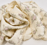 A cream-colored muslin baby blanket with a delicate pink and green floral print and lace trim, draped on a light neutral background.