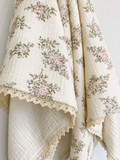 A cream-colored muslin blanket with a pink and green floral print and a scalloped lace hem, draped to show its texture and pattern.