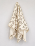 An off-white cotton throw blanket with a delicate cream floral pattern and scalloped edges, hanging against a plain light white wall.