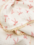 A close-up of a white muslin blanket with a pink bow print and a beige lace trim, folded to show its texture and pattern.