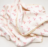 A white muslin baby blanket with a pink ribbon print and a beige lace trim, draped on a white background.