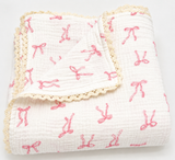 A folded white muslin baby blanket with a pink bow pattern and cream lace trim, shown on a clean white background.