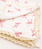 A folded white muslin blanket with a pink bow print and a cream-coloured lace trim, shown on a clean white background.