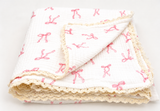 A folded white muslin blanket with a pink bow pattern and cream lace trim, displayed on a clean white background.
