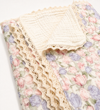 A folded muslin blanket with a cream-colored reverse side and a floral front print featuring pink and purple tulips with beige lace trim, shown on a white background.