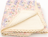 A folded cotton muslin blanket with a pastel pink and purple floral print on one side and a cream-colored reverse side, finished with a decorative cream lace border, shown on a white background.