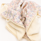 A folded cotton muslin baby blanket with a soft pink and lavender floral print on one side and a cream-colored reverse side with a delicate lace border, set against a plain white background.