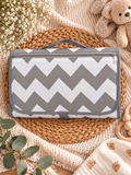 A gray and white chevron-patterned baby diaper change pad or storage pouch, styled in a cozy flat-lay setting on a woven placemat with baby props including a teddy bear, eucalyptus leaves, and a knit blanket.