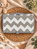 A gray and white chevron-patterned diaper bag or changing mat with a top handle, styled on a woven round mat with baby props including a teddy bear, knit blanket, and wooden beads.