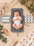 A baby lies on a gray mesh changing pad with attached sensory panels, photographed from above on a woven beige blanket with toys and flowers around it.