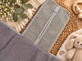A close-up styled product shot of a gray mesh change pad with a zipper, laid on top of a dark gray polka-dot fabric, surrounded by woven placemat, flowers, eucalyptus leaves, and a teddy bear.