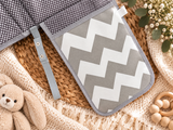 A baby change pad styled in a flat lay with a gray and white chevron patterned section, a black and white polka dot section, a woven basket, a cream knit blanket, a teddy bear, and baby toys.