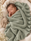 A sleeping newborn baby wrapped in a sage green knitted blanket with a ruffled border, resting in a woven basket next to a beige plush bunny toy.